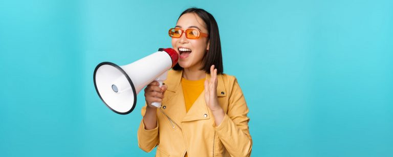 stylish-asian-girl-making-announcement-megaphone-shouting-with-speakerphone-smiling-inviting-people-recruiting-standing-blue-background